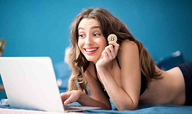 Young and happy woman buying online with cryptocurrency concept, woman holding golden crypto coin lying with laptop on the bed at home